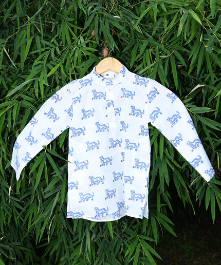 A long-sleeved white kurta with blue block print tiger patterns hanging on a hanger against a backdrop of green foliage.