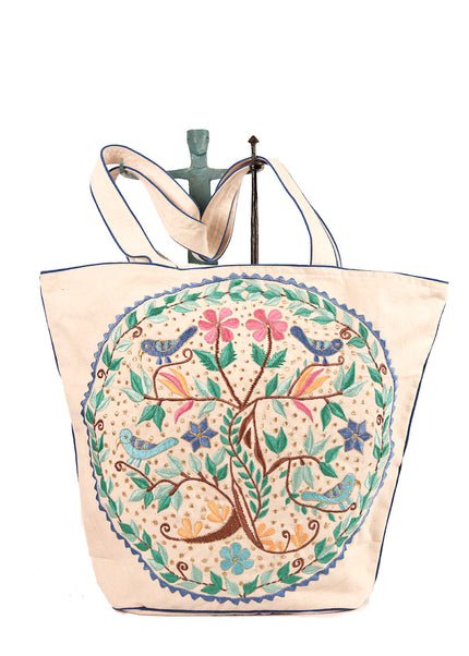A beige cotton tote bag with a 'Tree of Life' inspired embroidery on one side and a paisley motif on the other, featuring a handle and a branded tag on top.