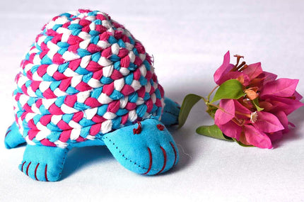 A stuffed toy resembling a tortoise with blue and pink patterned shell, resting against a background of pink flowers.
