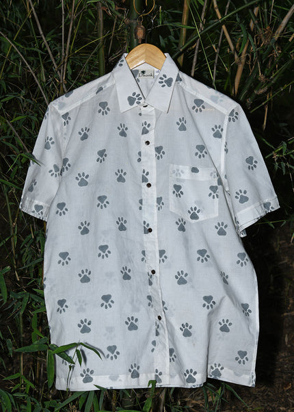 Front view of a children's short-sleeved white button-down shirt featuring a repeating blue paw print pattern, hanging on a wooden hanger against a background of green bamboo leaves.