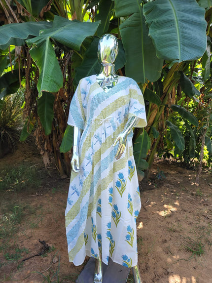 A mannequin showcasing a sleeveless kaftan with blue and green stripes and bird patterns, standing in a tropical setting with banana plants in the background.
