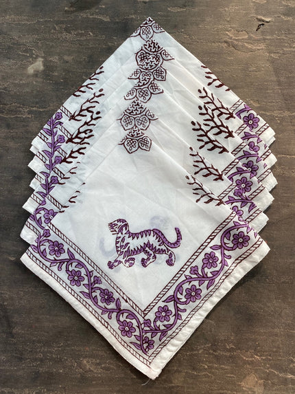 A set of folded cloth napkins with a hand block printed purple animal pattern on a white background, displayed on a wooden surface.