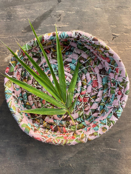 A round basket with a multicolored pattern, containing some greenery, placed on a grey surface.