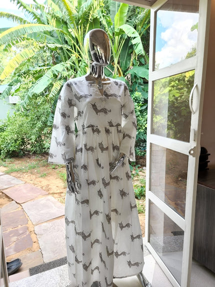 A maxi dress displayed on a mannequin, featuring a white color with a hand block print pattern, standing in a room with a tropical landscape visible through the window.