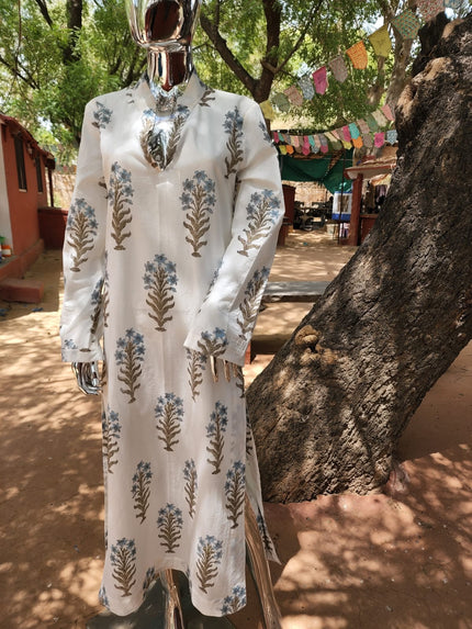 A white kaftan with blue and green floral hand block print, displayed on a mannequin outdoors with a tree and traditional buildings in the background.